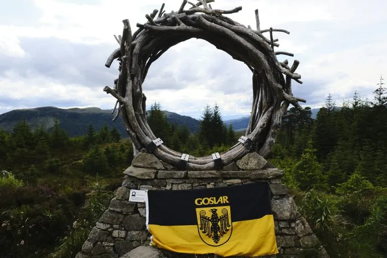 Goslar-Fahne an einem Steinsockel mit Ring aus Treibholz, dahinter bewaldete Hügel in Schottland